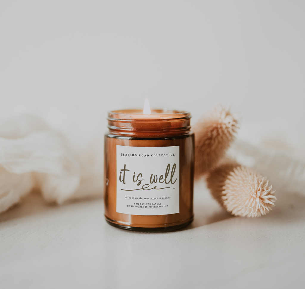 Candle in a amber jar with 'it is well' label on a neutral background