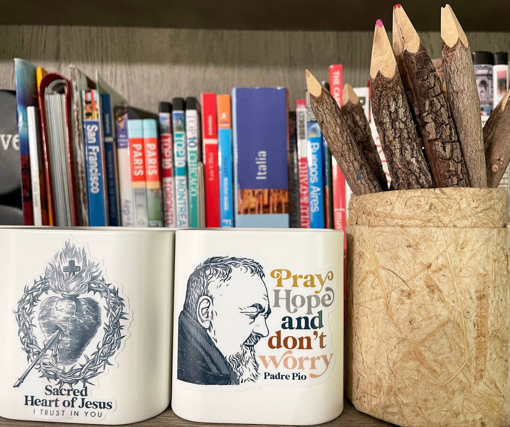 Two white ceramic mugs displayed on a shelf in front of a bookcase. The left mug is white with a Sacrad Heart of Jesus art illustration of a heart and the words 'Sacred Heart of Jesus I TRUST IN YOU'. Right mug with Saint Padre Pio 'Pray hope and don't worry.