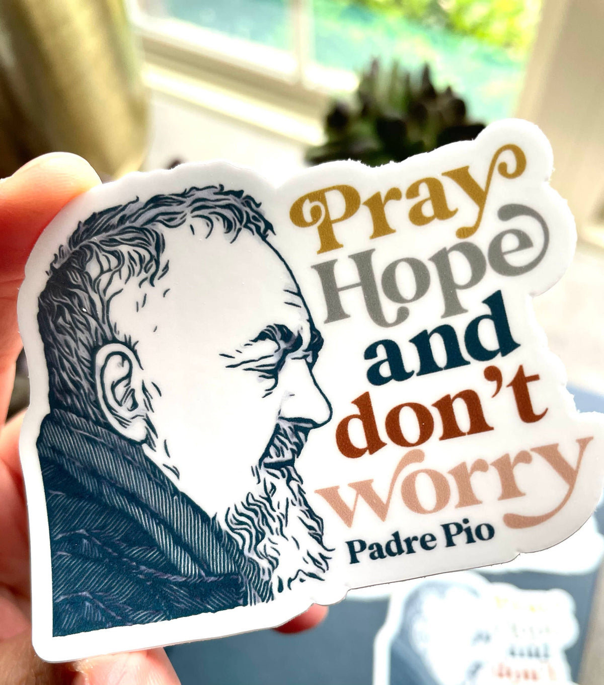 A hand holds a die-cut sticker featuring a black-and-white illustrated portrait of Saint Padre Pio alongside the text 'Pray Hope and don't worry' in stylized brown, gray, blue, and orange lettering.