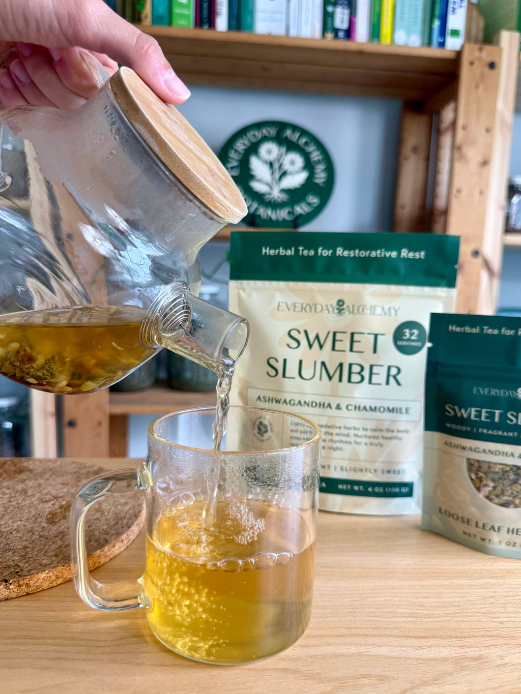 A hand pours golden herbal tea from a glass teapot with a bamboo lid into a clear glass mug. Two bags of Everyday Alchemy Sweet Slumber herbal tea are displayed on a wooden table in front of a bookshelf.