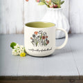 White mug with floral design and text on a wooden surface with flowers and a vase in the background
