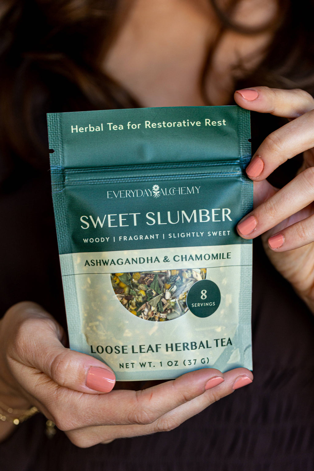 A person holds a stand-up pouch of Everyday Alchemy Sweet Slumber loose leaf herbal tea. The packaging is dark teal green with white text and a circular window showing the loose leaf tea inside.