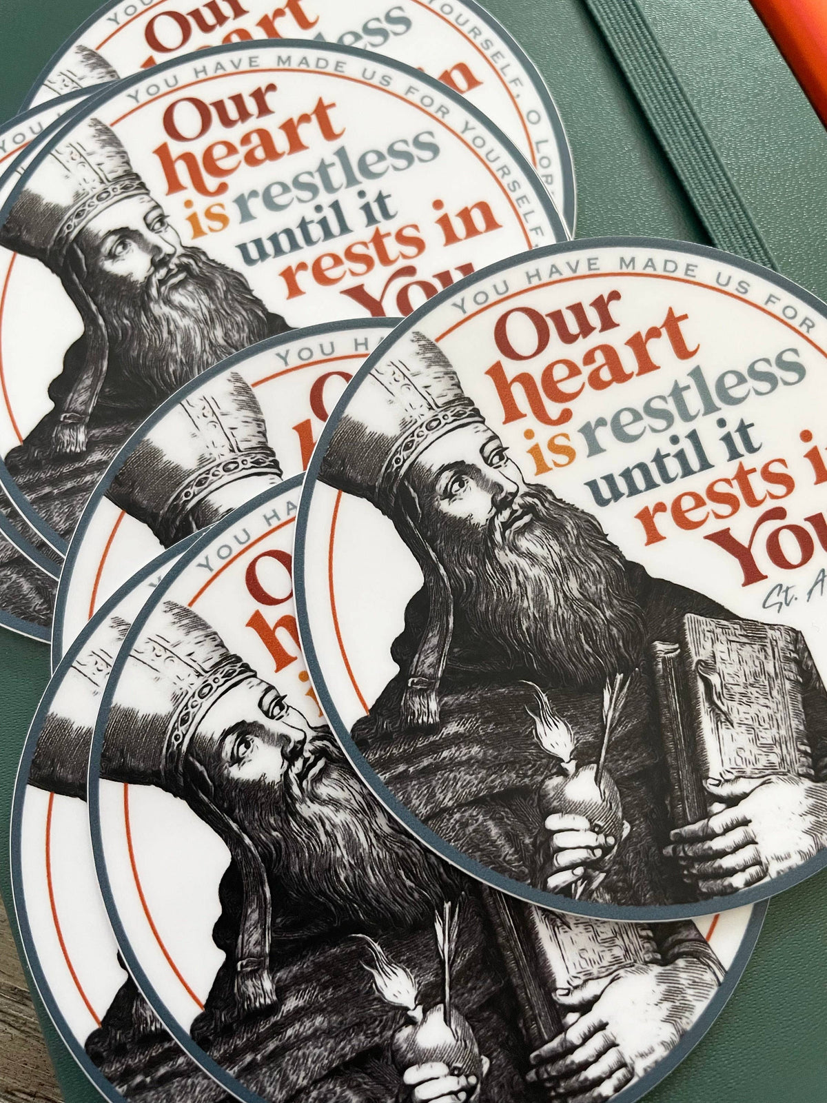 Several overlapping oval stickers or decals with a black-and-white illustration of a bearded Saint Augustine and the text 'Our heart is restless until it rests in You,' arranged on a green surface.
