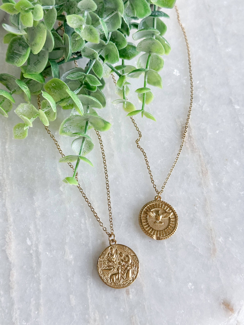 Two gold coin pendant necklaces with detailed religious engravings, displayed on a light marble surface next to green eucalyptus leaves.