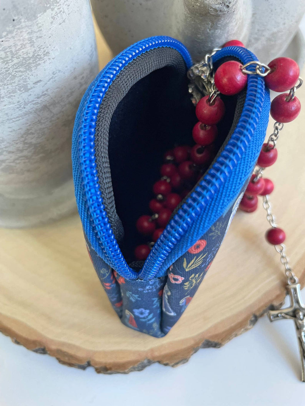 Blue zippered pouch with floral pattern containing red rosary beads and a cross, on a wooden surface.