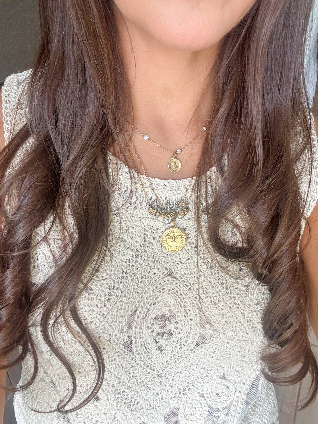 A close-up of a woman wearing layered gold necklaces, including a prominent round gold pendant with a sunburst design and a smaller charm necklace, over a white lace top.