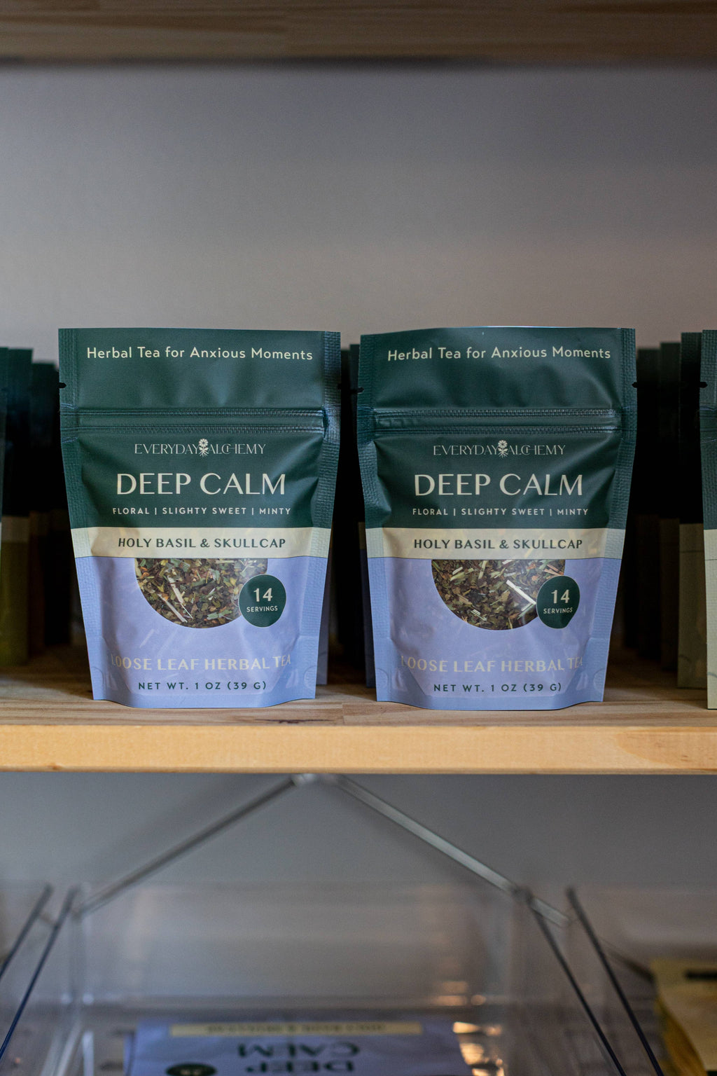 Two stand-up pouches of Everyday Alchemy Deep Calm Holy Basil & Skullcap herbal tea displayed on a wooden shelf.