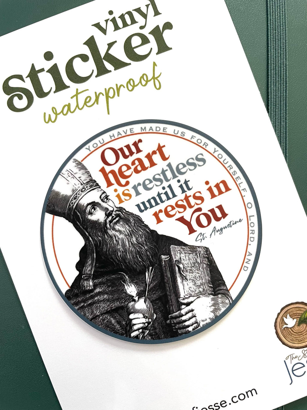 A circular vinyl sticker shown on its white backing card, with a dark green textured surface in the background. The sticker features a black and white illustration of Saint Augustine holding a book, surrounded by the quote 'Our heart is restless until it rests in You  