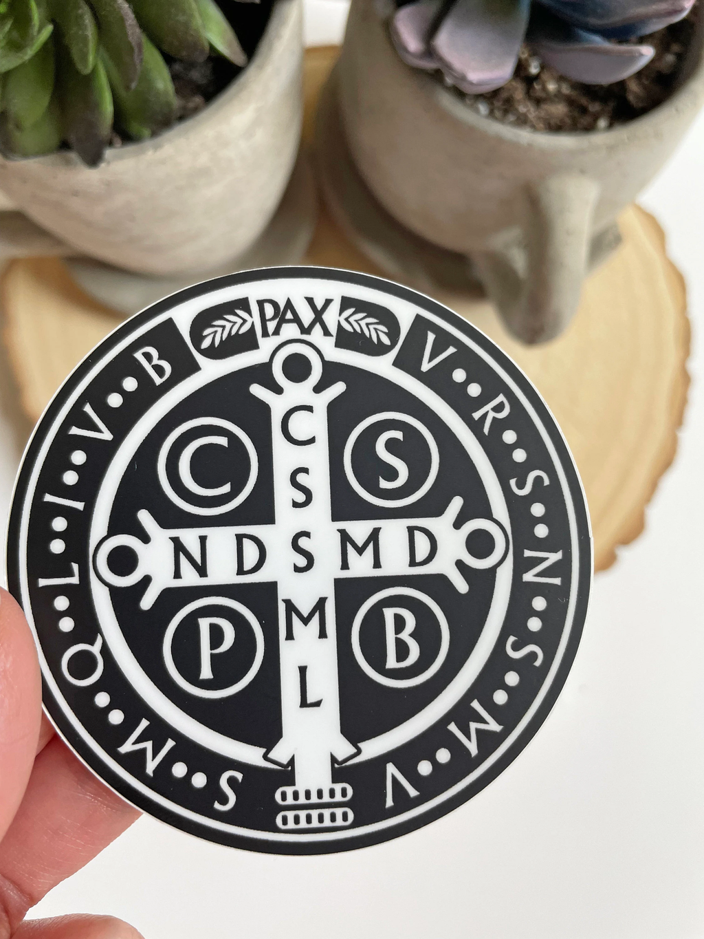 Circular black and white emblem with religious symbols held by a hand, with plants in the background.St. Benedict Medal Waterproof Sticker
