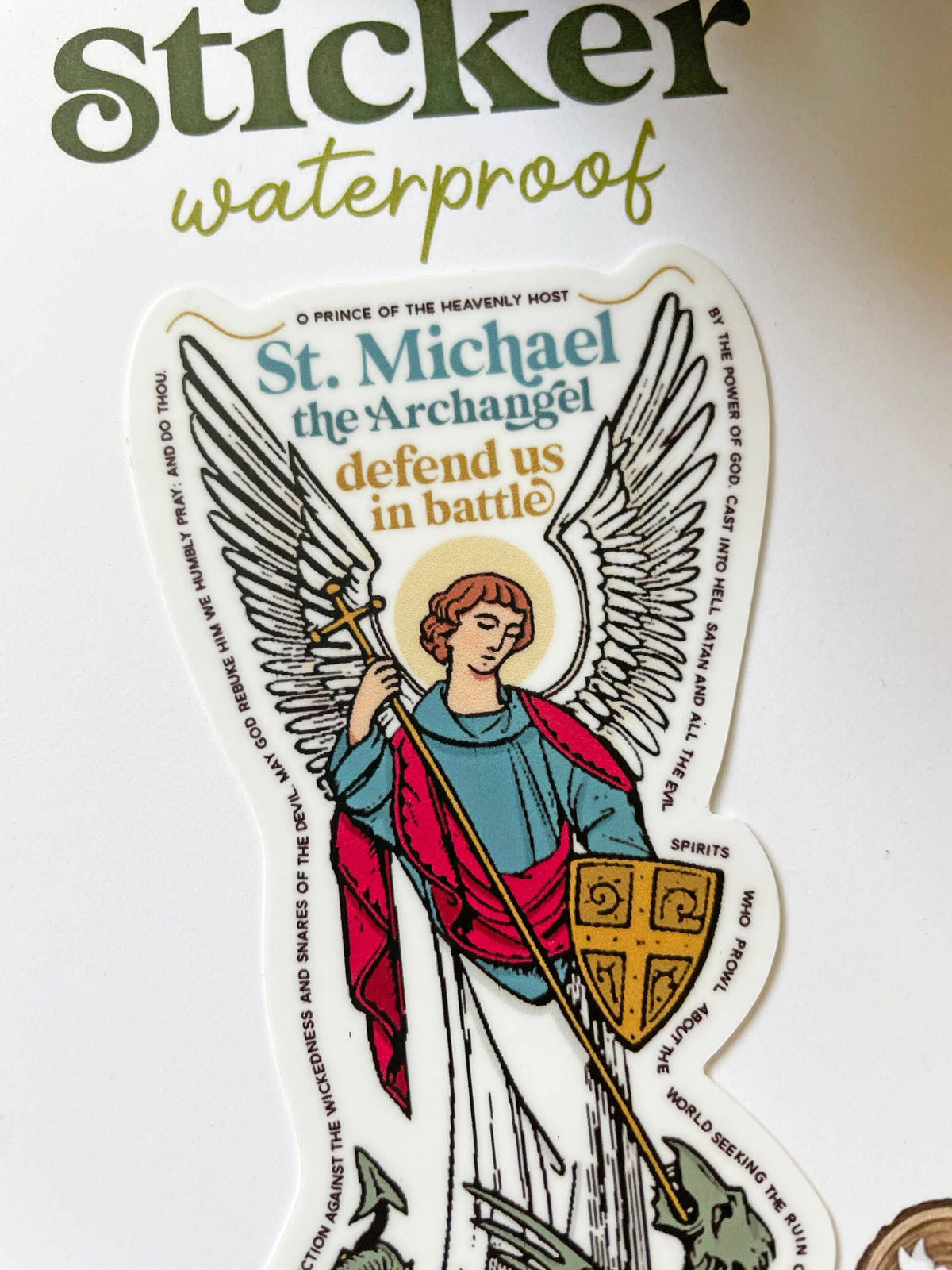 A die-cut sticker featuring an illustration of St. Michael the Archangel with wings, a sword, and shield