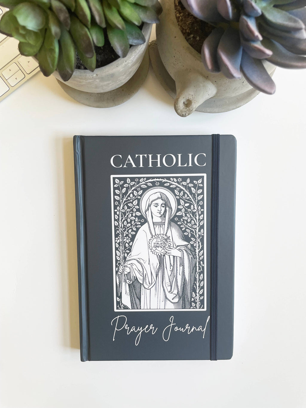 Catholic prayer journal with a decorative cover on a white surface with plants.