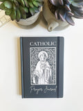 Catholic prayer journal with a decorative cover on a white surface with plants.