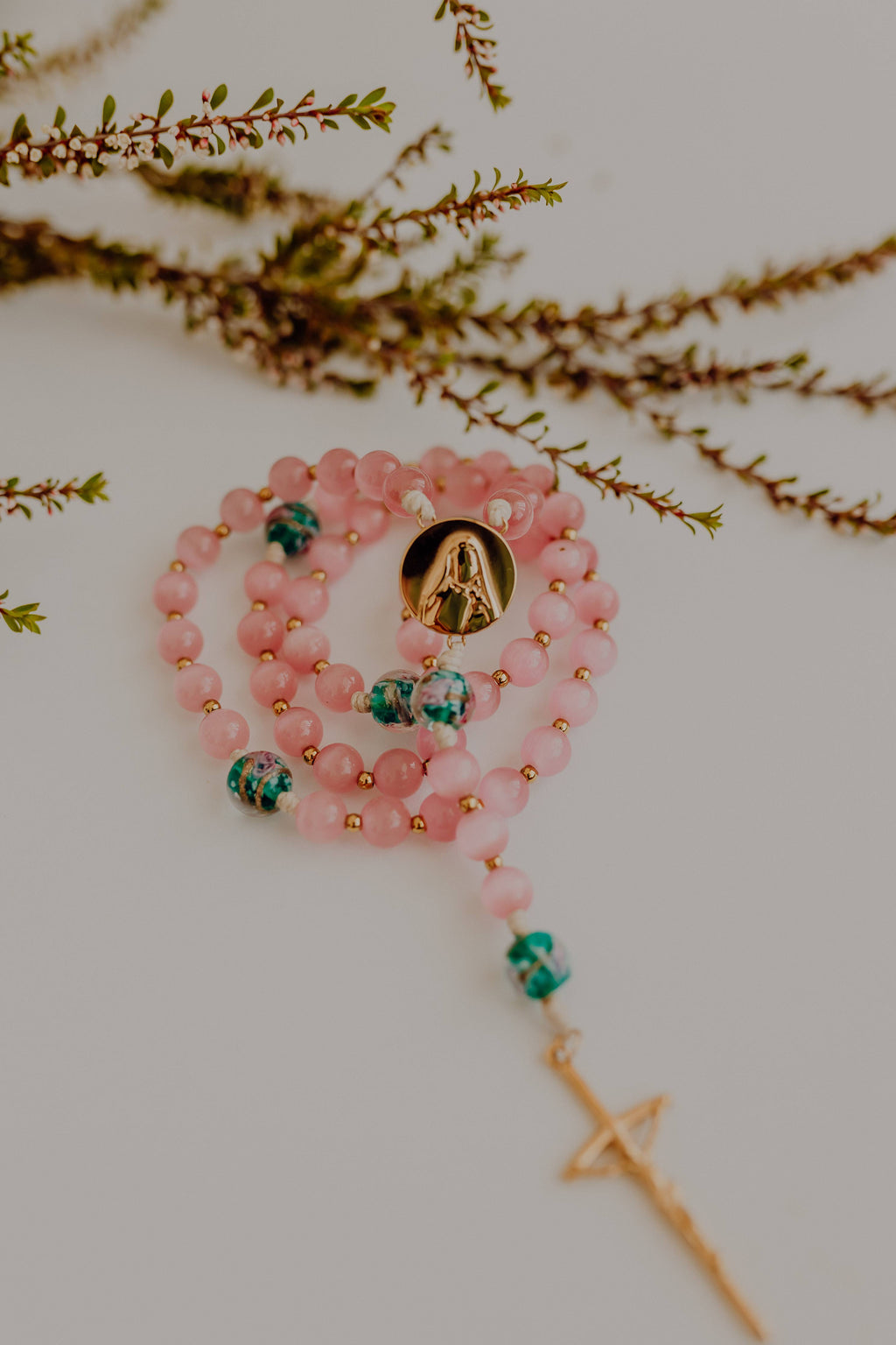 Pink beaded Rosary with green accents on a white surface with branches in the background