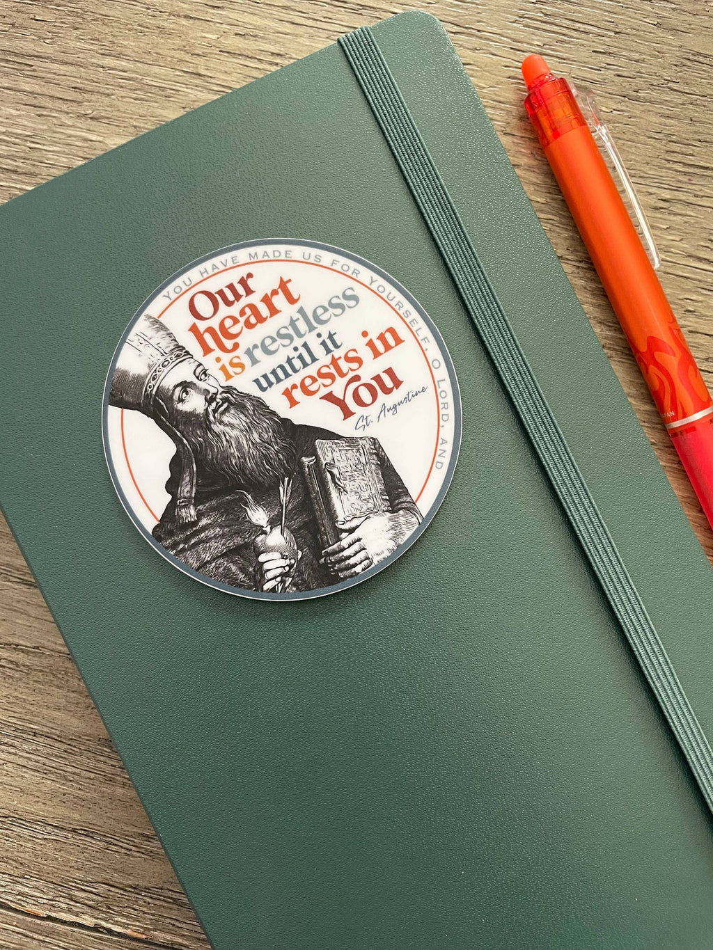 A dark green hardcover notebook with an elastic closure strap, featuring a circular printed sticker with a black and white portrait of Saint Augustine and the quote 'Our heart is restless until it rests in You'. An orange pen rests on the right side on a wooden surface.