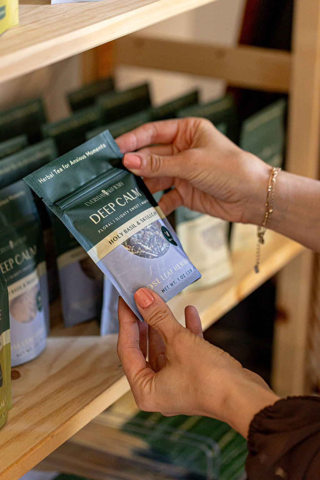 A person holding a single packet of Everyday Alchemy Deep Calm tea on a light wood shelf.