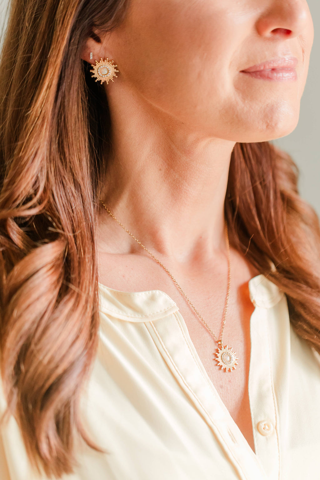 Close-up of a woman wearing a delicate gold necklace with a sunburst pendant and matching sunburst stud earrings, paired with a cream-colored blouse.