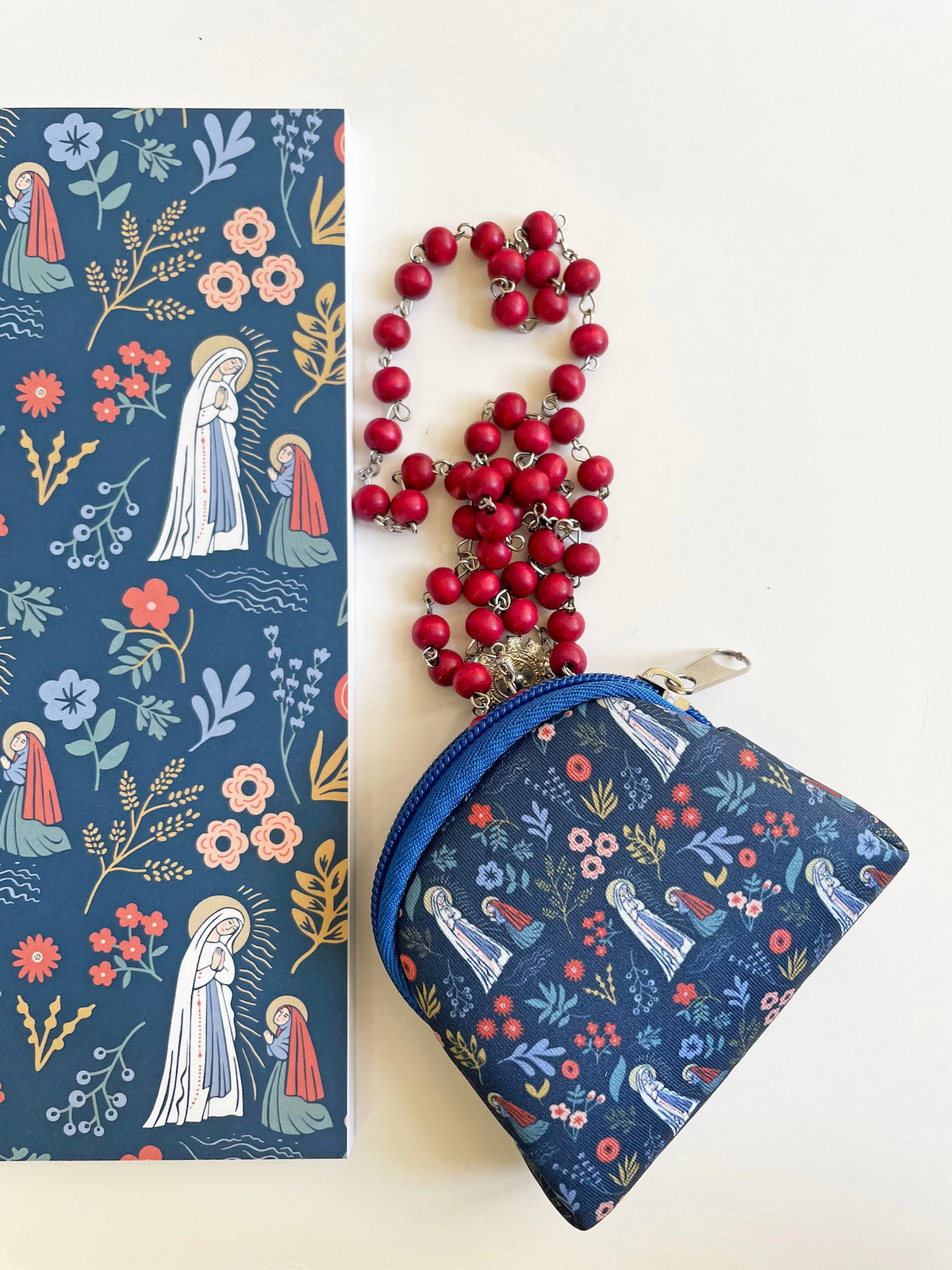 A navy blue zippered pouch with a floral and religious print, shown next to a matching printed card or booklet and a red beaded rosary necklace with silver links. The background is a clean off-white surface.
