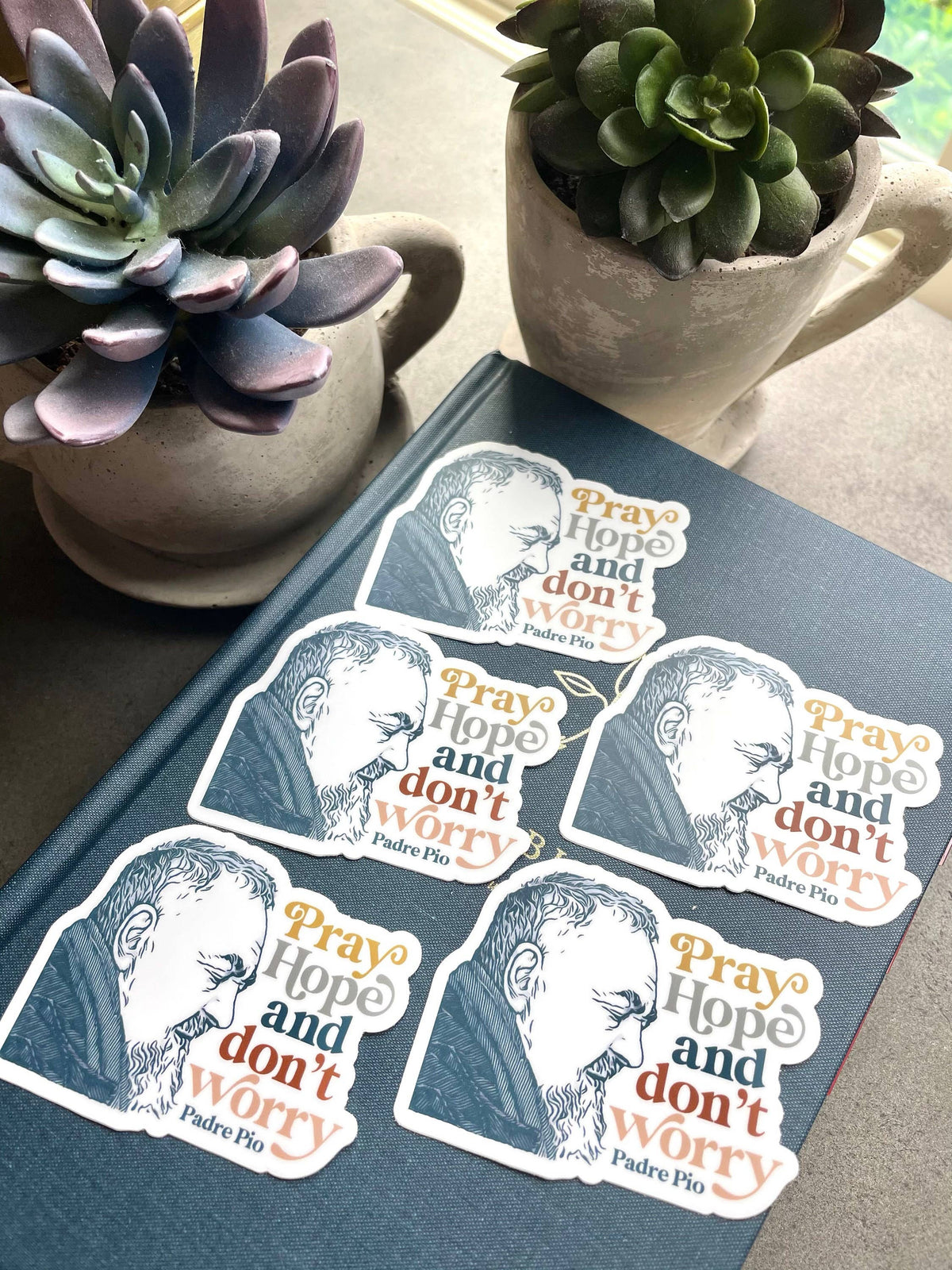 Five die-cut stickers featuring a stylized portrait of Saint Padre Pio with the words 'Pray Hope and don't worry' in orange, blue, and white, arranged on a dark book with two potted succulent plants in the background.