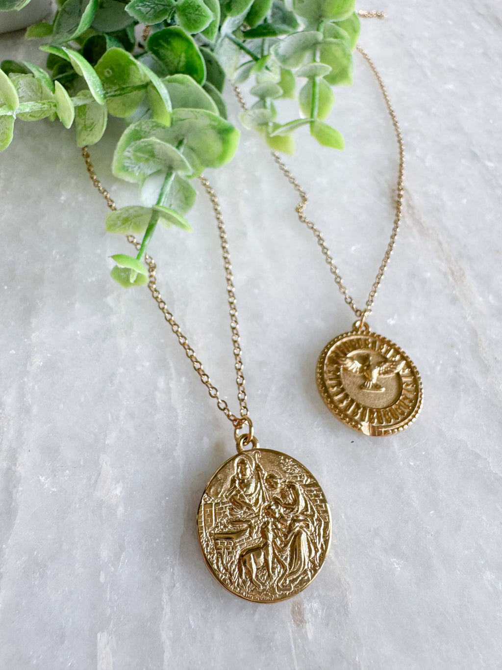 Two gold coin pendant necklaces displayed on a light marble surface with green eucalyptus leaves. One pendant shows a detailed religious figure, and the other shows a sun and moon symbol.