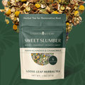A stand-up pouch of Everyday Alchemy Sweet Slumber loose leaf herbal tea, shown against a dark green background with loose dried tea leaves arranged around the top and sides. The pouch is dark teal on the upper half and cream on the lower half, with product text and a small circular photo of mixed herbs.