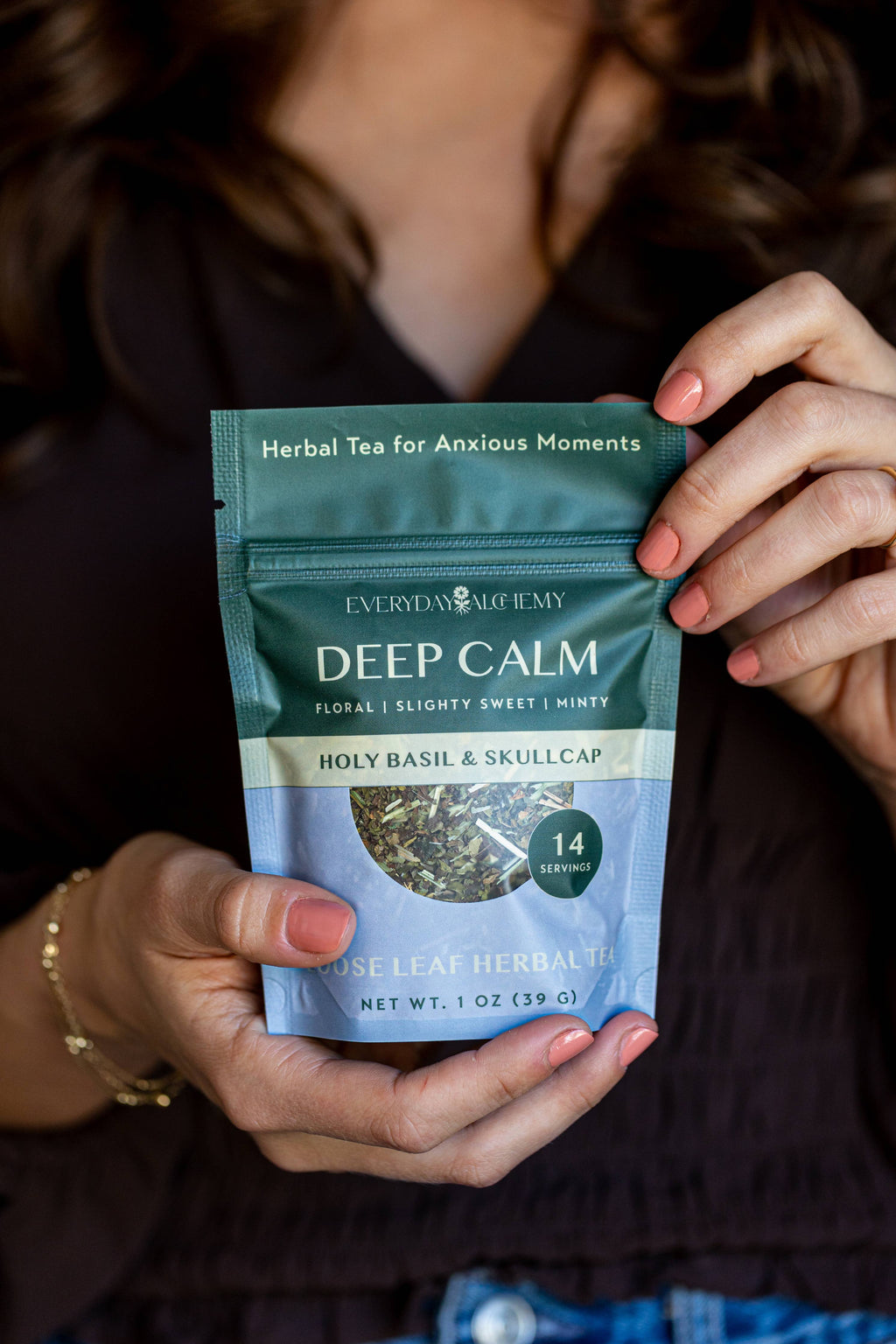 A person holds a stand-up pouch of Everyday Alchemy Deep Calm herbal tea. The packaging is dark teal green with white and light blue text and a circular window showing loose leaf tea inside.