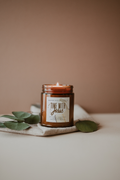 Candle in a glass jar with a label "Time with Jesus" on a beige surface with green leaves