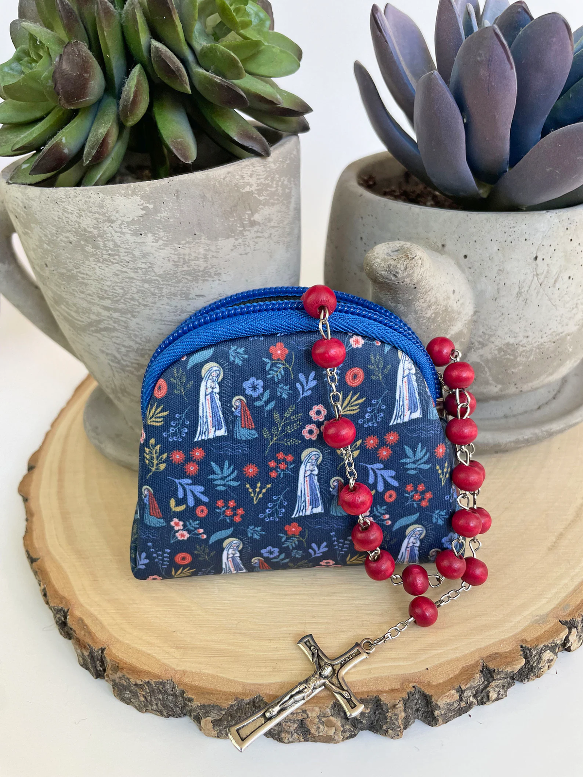Blue floral-patterned rosary pouch with red rosary beads on a wooden surface with succulents in the background
