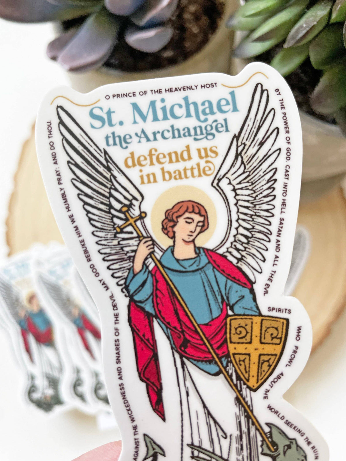 A die-cut sticker featuring an illustration of St. Michael the Archangel with large white wings, a blue tunic, and a red cloak, holding a gold cross and shield.